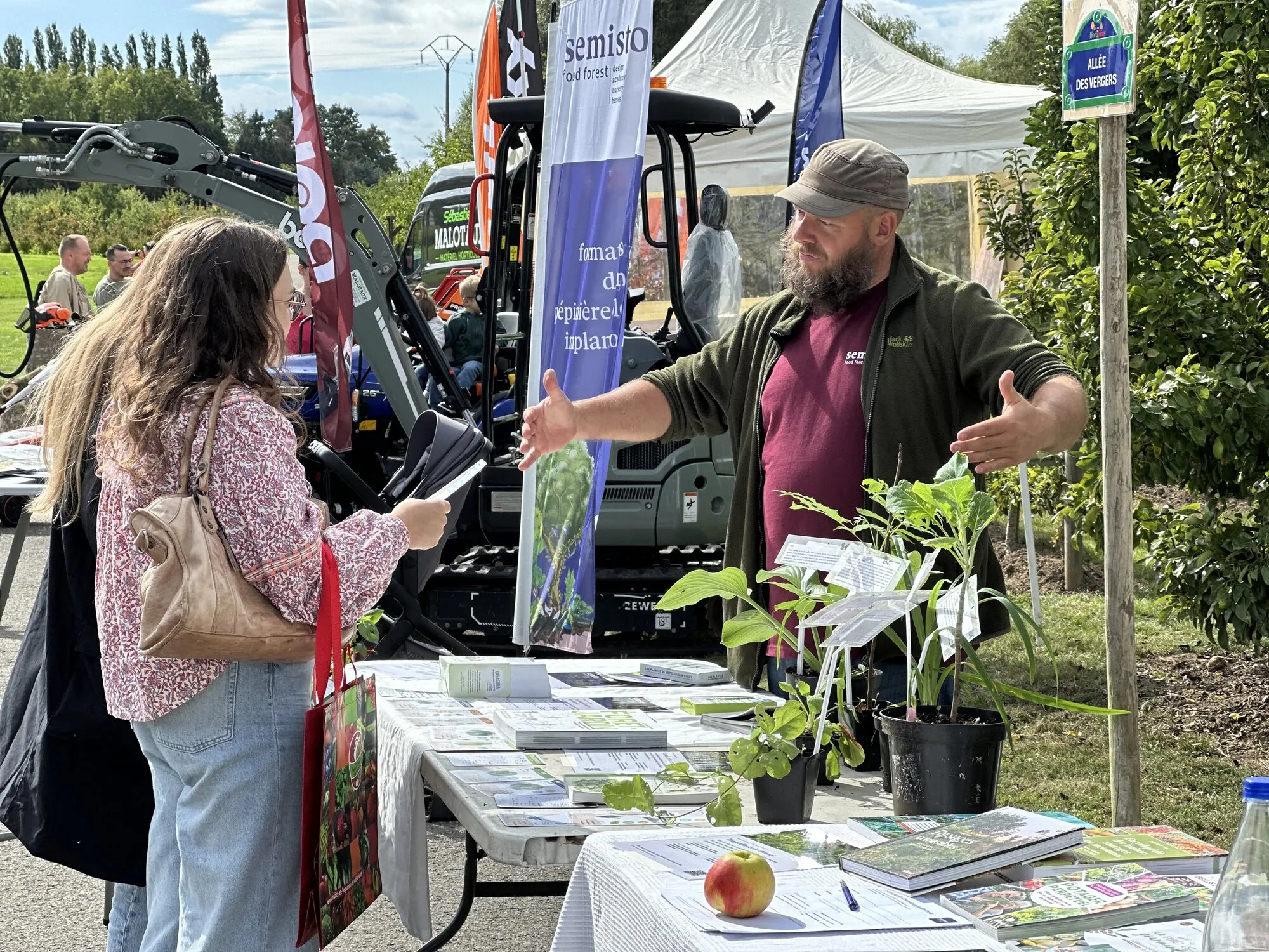 Sur le stand Semisto aux Hortifolies 2026 : un membre du collectif explique le jardin-forêt à une visiteuse, entre plants, livres et brochures